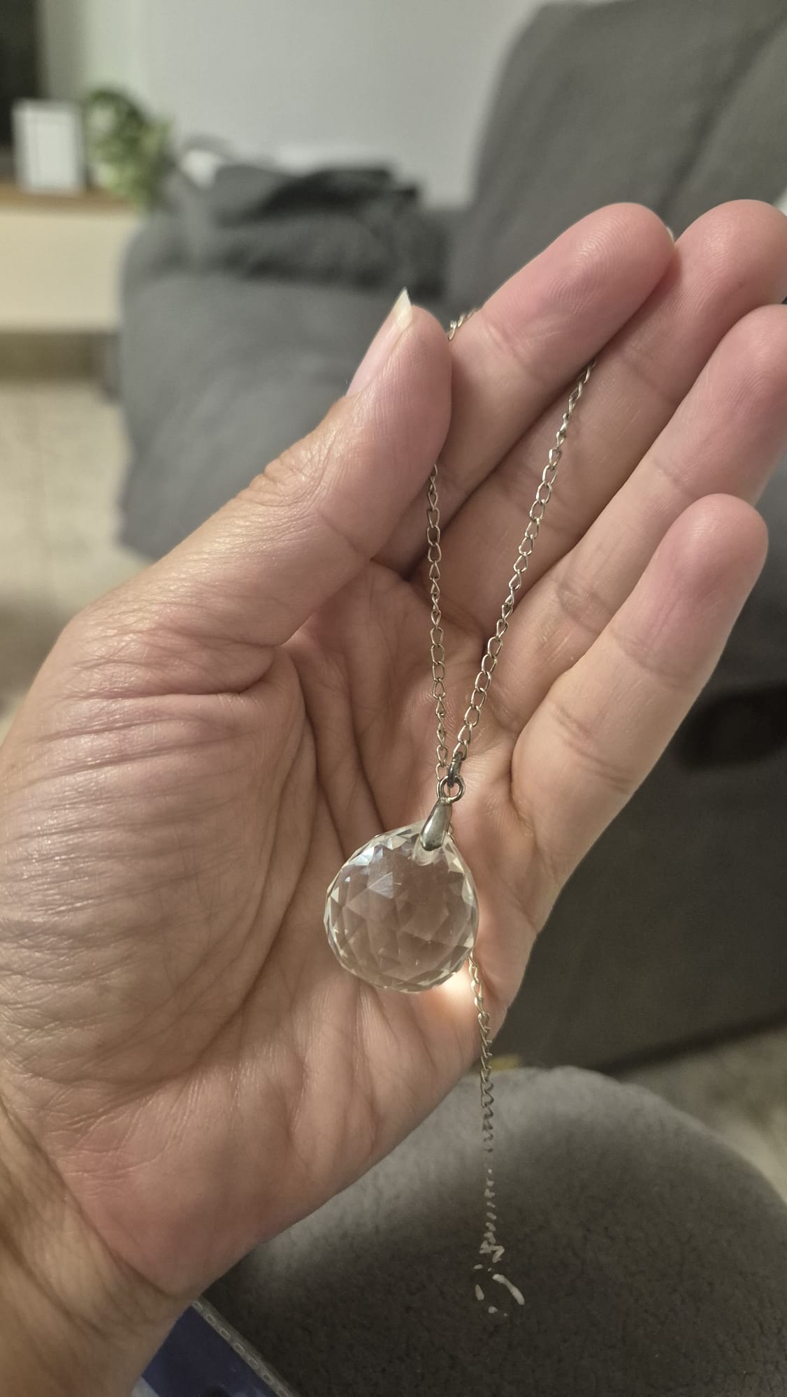 יד מחזיקה במטוטלת זכוכית שקופה עם פיסול גאומטרי, תלויה על שרשרת מתכתית דקה