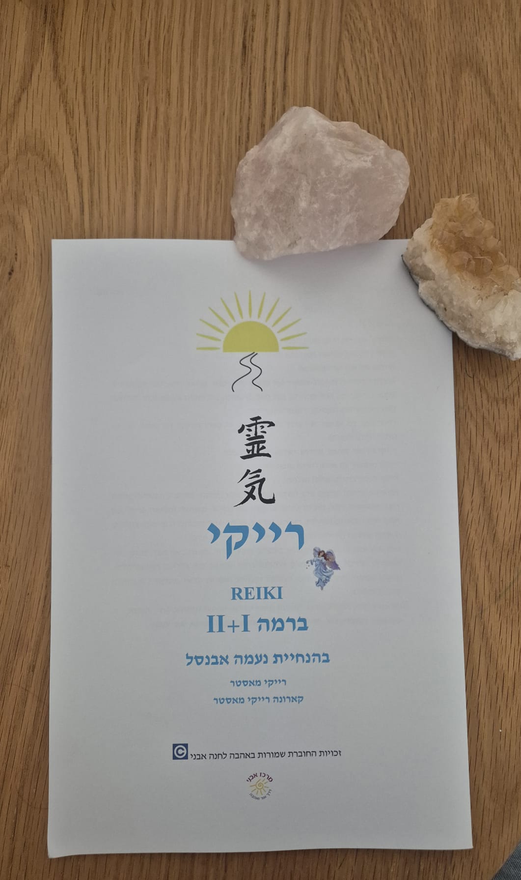 דף מידע על סדנת רייקי, עם כותרת "רייקי II+I" ולוגו של שמש. מסמכים מונחים על שולחן עץ, עם אבני קריסטל בצדדים
