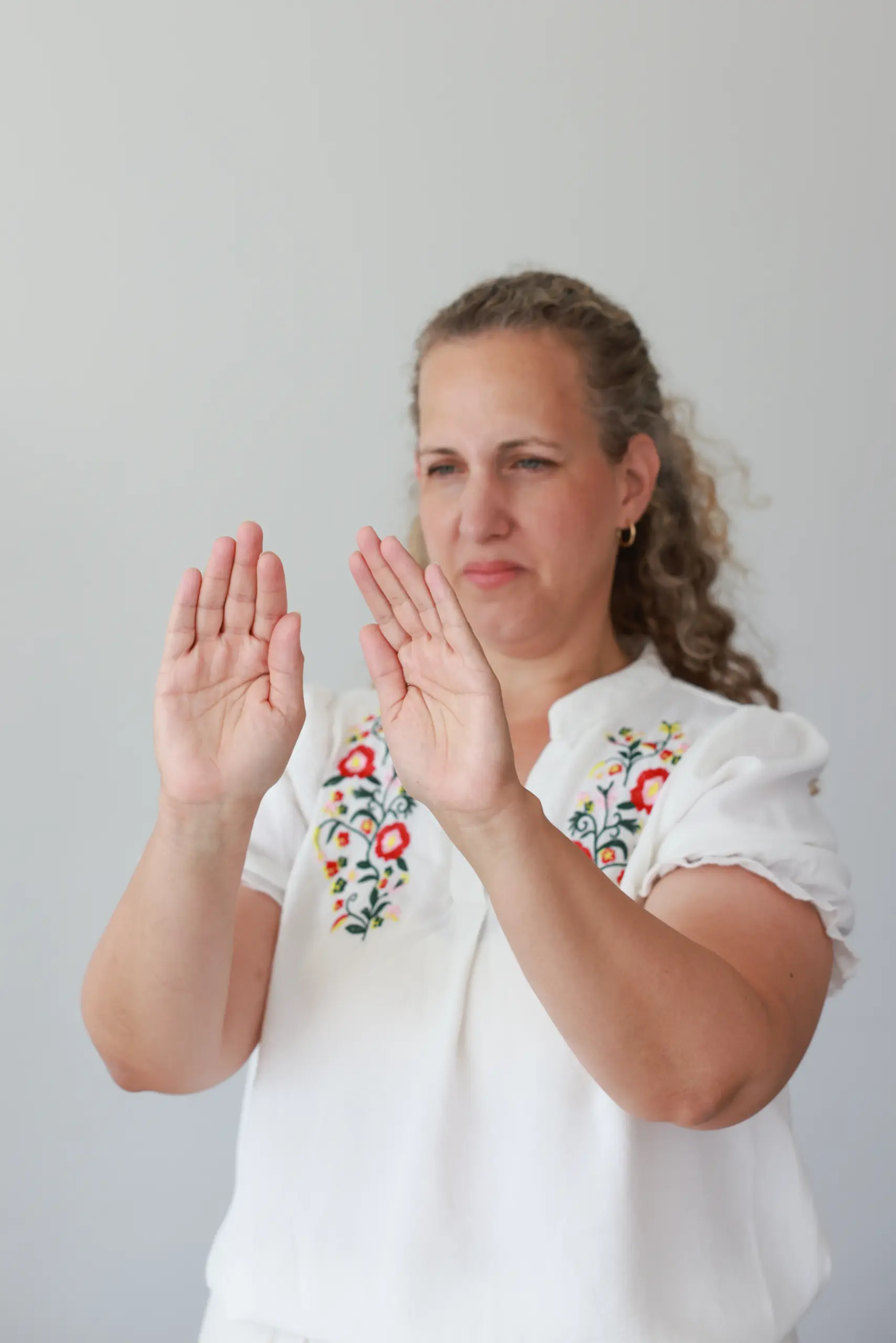 אישה עם שיער גלי, לבושה בחולצה לבנה עם רקמה צבעונית, מבצעת תנועות ידיים הממחישות טיפול מרחוק. הרקע בהיר ופשוט